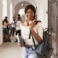African girl with backpack and notes standing on corridor of university and smiling at camera. Pretty student girl with headphones near neck going to class room. Concept of studying.